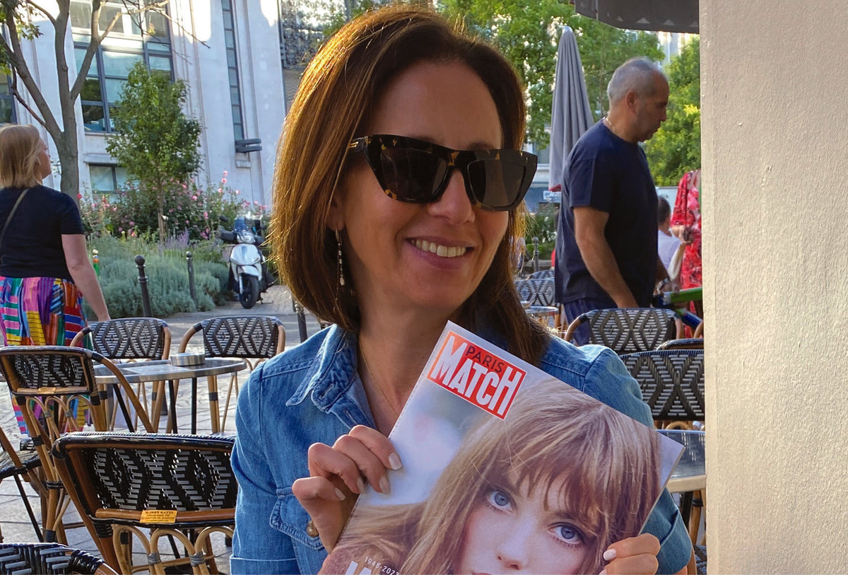 Natalie smiling at a Paris café table, holding up a copy of Paris Match